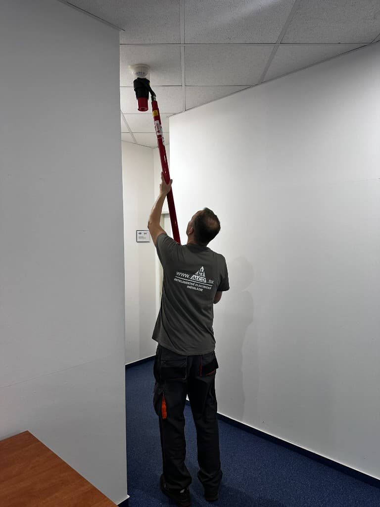 Technik ALCO.SK pri skúške dymového detektora na strope – elektrická požiarna signalizácia (EPS), červená skúšobná tyč, logo ALCO.SK na tričku, inteligentné elektrické inštalácie