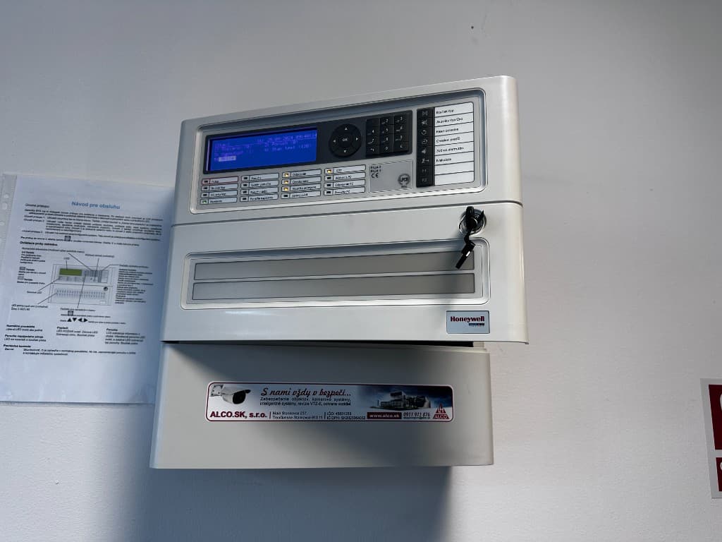Požiarna ústredňa Honeywell – modrý displej, stav požiarov a porúch, numerická klávesnica, návod na obsluhu na stene. Nálepka ALCO.SK – elektrická požiarna signalizácia EPS, montáž a servis.