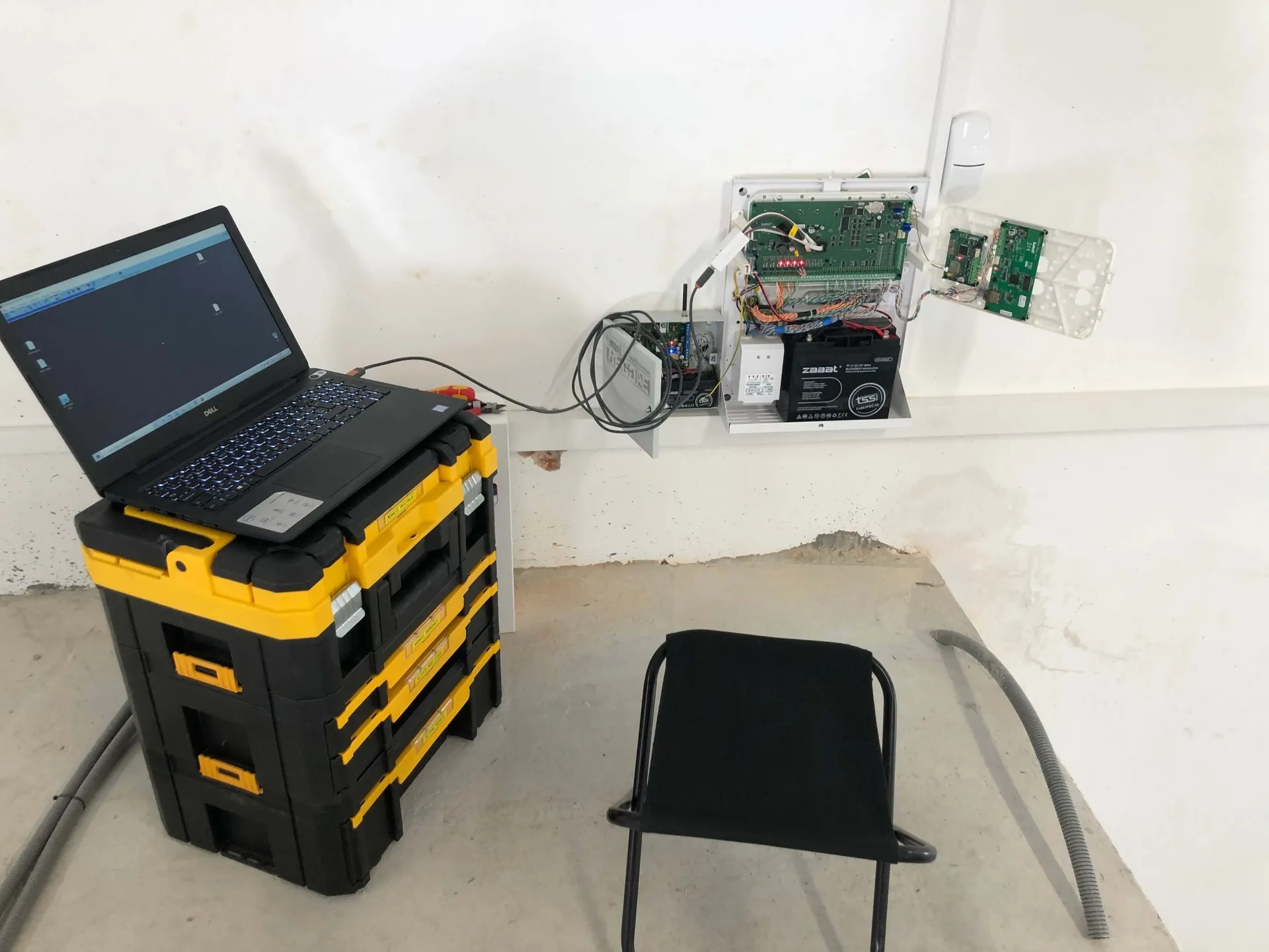 Inštalácia a servis zabezpečovacieho systému ALCO – otvorená ústredňa na stene, obvodová doska, batéria, pripojený laptop a pracovné prostredie. Servis alarmov a EZS.