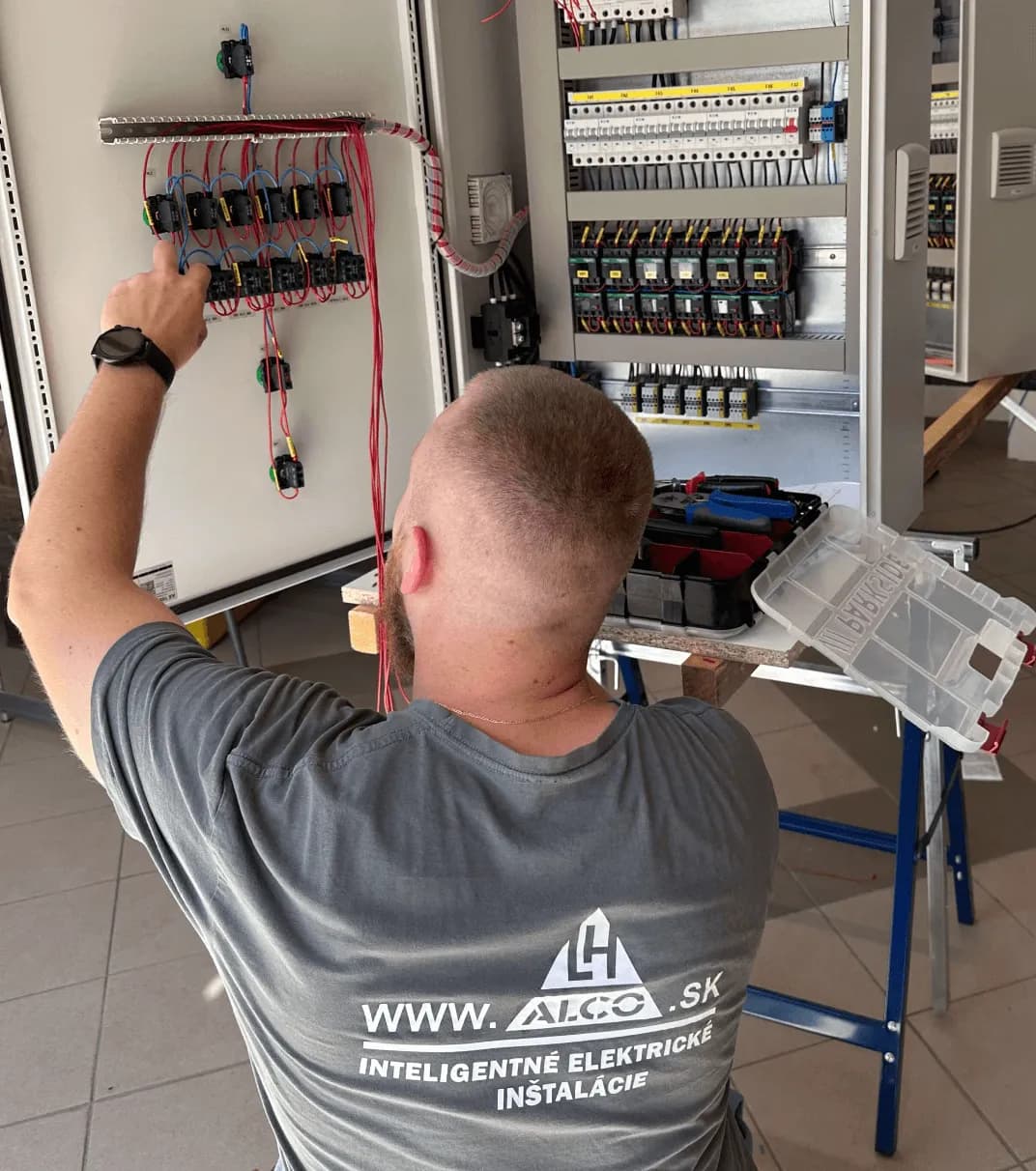 Elektrikár ALCO.SK s logom na tričku pri práci na riadiacom paneli rozvádzača NN – inteligentné elektrické inštalácie